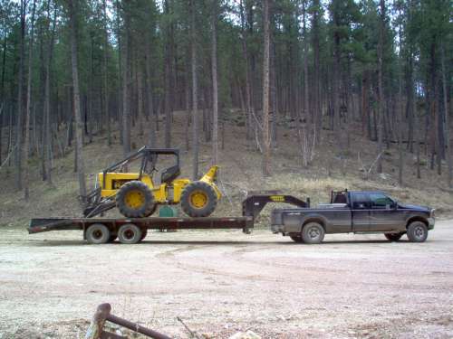 Yellow tractor on flatbed trailer towed by dark pickup truck in wooded area