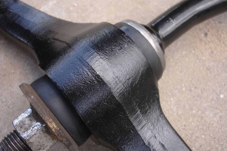 Black painted automotive suspension component with a rubber bushing and a bolt. The part has visible casting marks
