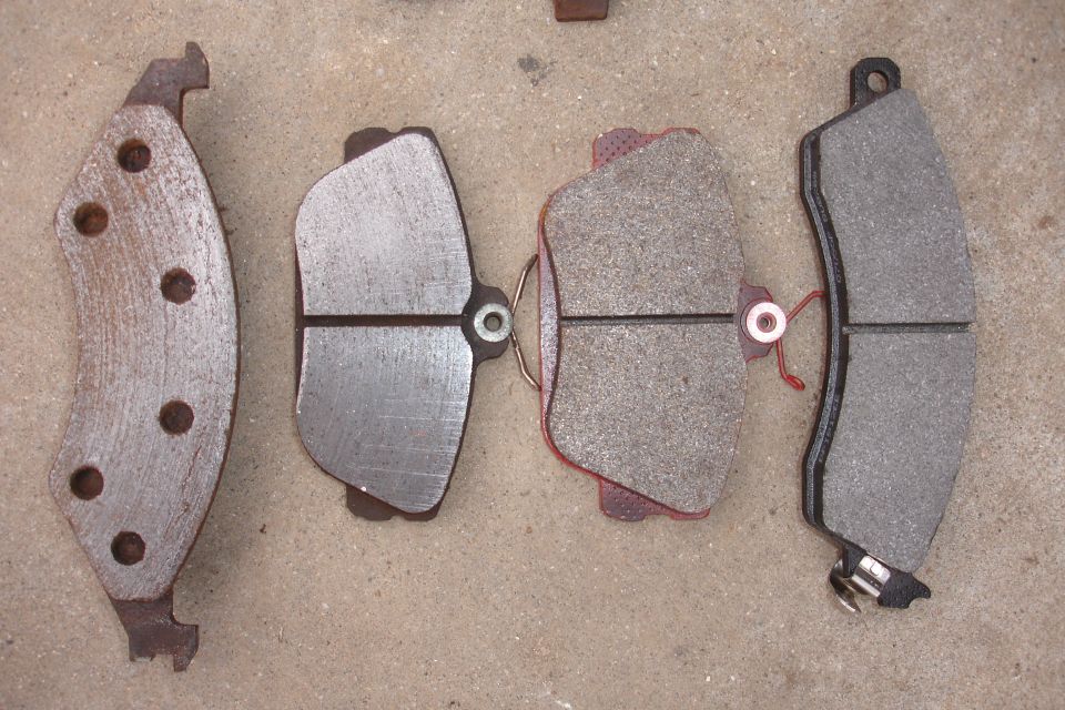 Four brake pads on a concrete surface. Two are new with grey friction material, one is worn with red backing, and one is old and rusty