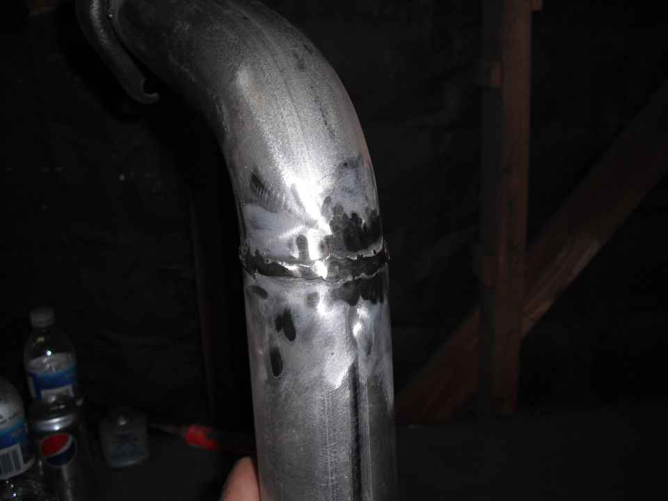 Close-up of a metal exhaust pipe with a visible weld seam. Bottles and a can are in the background