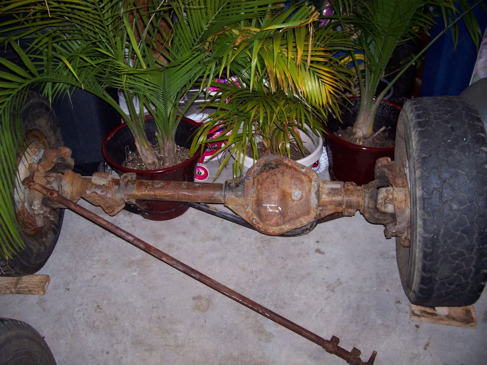 Rusty solid front axle with differential and wheels on wood blocks in a garage, palm plants behind