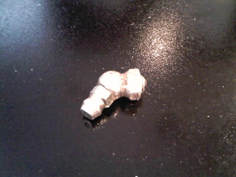 Broken-off metal zerk grease fitting lying on a dark oily surface