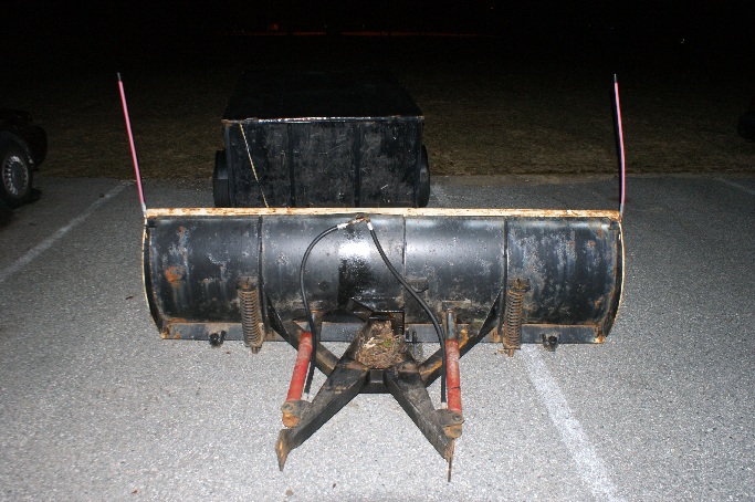Front view of a snow plow attachment with hydraulic lines and springs. Two pink markers are on top