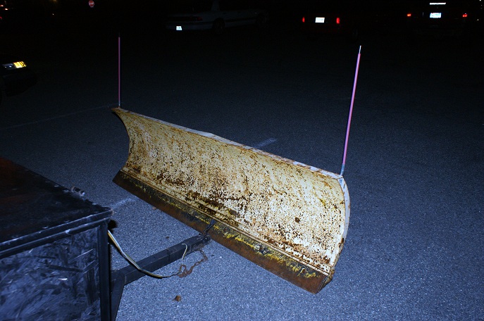 Rusty yellow snow plow with pink markers attached to a trailer hitch