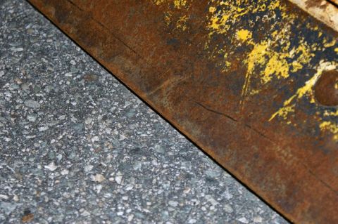 1979 Ford Bronco - Close-up of a heavily rusted metal surface with chipped yellow paint, resting on asphalt.