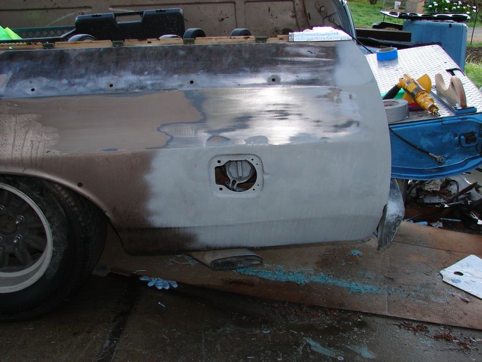 Rear quarter panel of a vehicle undergoing bodywork, showing primer and sanding marks. Fuel filler door is open