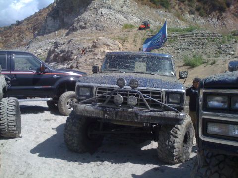 1979 Ford Bronco - Several off-road trucks covered in mud are parked on a sandy, rocky terrain.