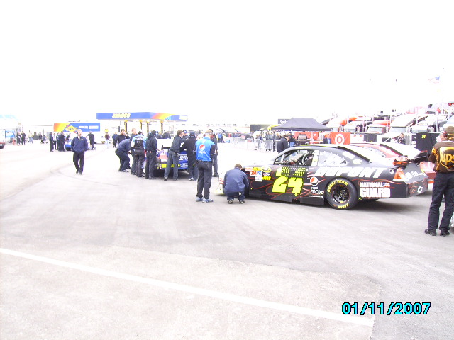 Race cars with crew members working on them, pit area with gas station in background