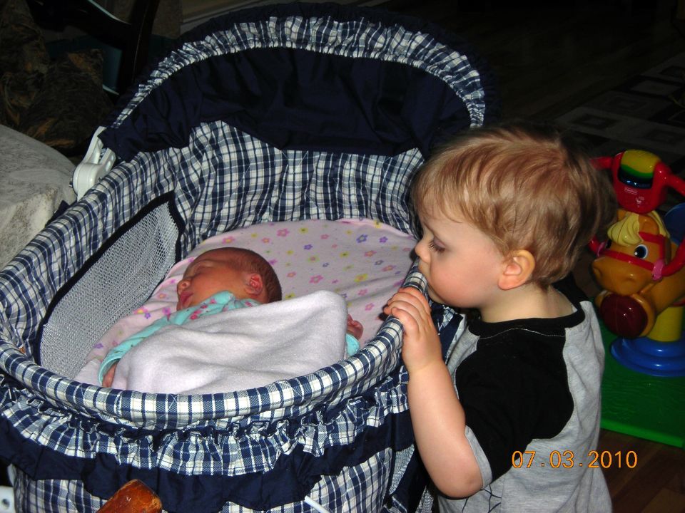 Toddler peers into a bassinet at a sleeping baby. Date stamp reads 07.03.2010
