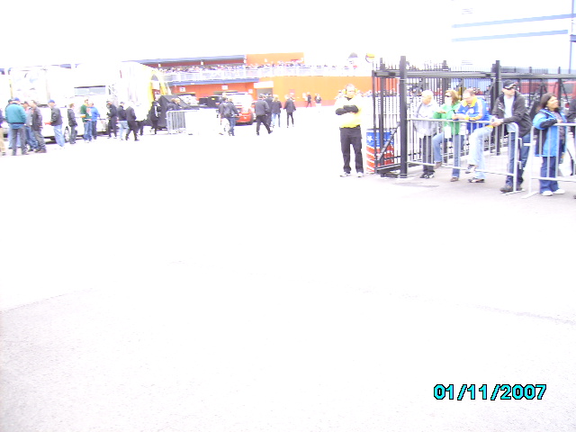 Crowd gathered near a gate with vehicles and stadium in background