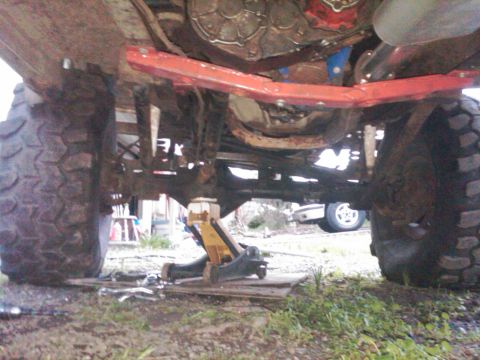 1978 Ford Bronco - The underside of a vehicle is shown, large off-road tires, a jack, and a glimpse of another vehicle in the distance.
