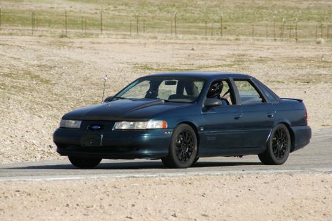 1995 Ford Taurus - A dark blue 1995 Ford Taurus is captured on a race track with a driver wearing a helmet.