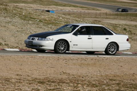 1995 Ford Taurus - White 1995 Ford Taurus with a black hood on a racetrack, seen from the side.