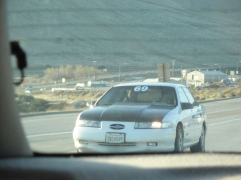 1995 Ford Taurus - A white 1995 Ford Taurus with a black hood and the number 69 on its windshield drives on a highway.