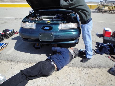 1995 Ford Taurus - Two people are working on the engine of a 1995 Ford Taurus that is raised on jack stands.