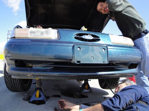 1995 Ford Taurus - Two men are working on the front of a teal Ford Taurus that is raised on jack stands.