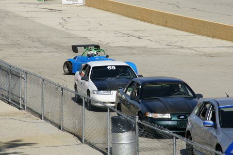 1995 Ford Taurus - A white Ford Taurus with a black hood is seen on a race track, with a blue race car in front.