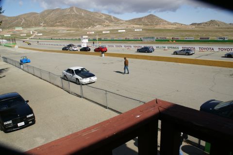 1995 Ford Taurus - A white sedan with a black hood is parked on the side of a race track, with other vehicles and spectators visible.