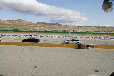 1995 Ford Taurus - Two cars are on a race track with a backdrop of hills and sponsor banners.