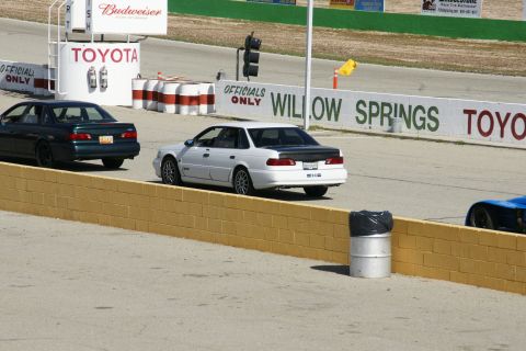 1995 Ford Taurus - A white 1995 Ford Taurus sedan is seen on a race track, with other vehicles and track signage in the background.