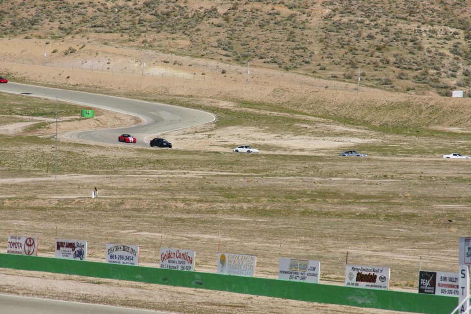 Cars on a winding race track with dry hills in background. Signs visible in foreground