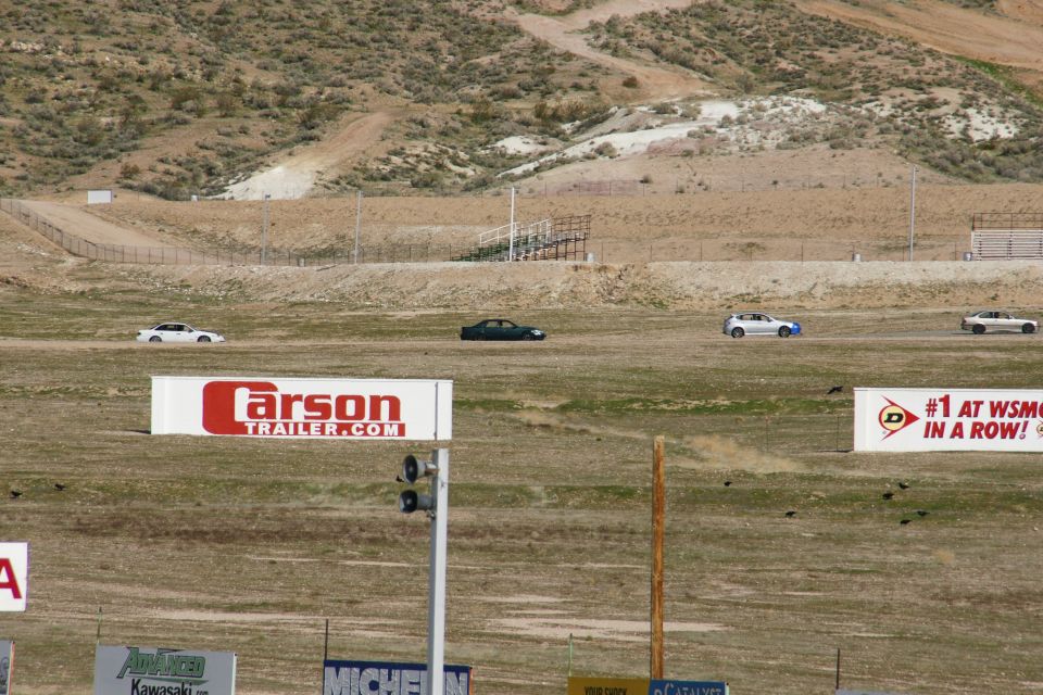 Four cars drive on a dry, grassy racetrack with hills in the background. A white car is in the foreground