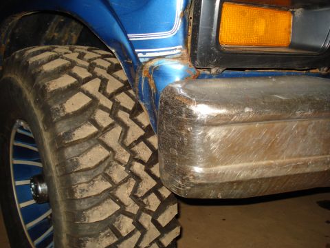 1985 Ford F-150 - Close-up of a blue truck's front corner, showing a large tire and worn chrome bumper.
