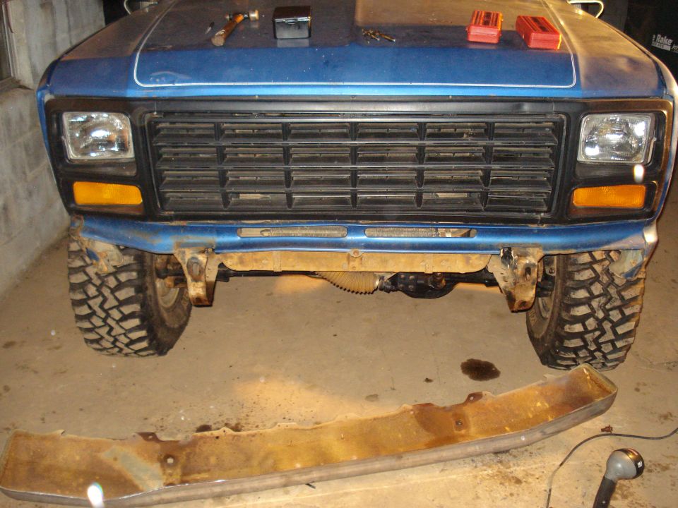 Blue pickup truck front end with a large grille and off-road tires. A rusty bumper lies on the ground