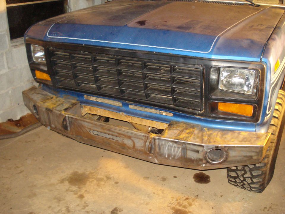 Blue pickup truck front end with a custom metal bumper and a black grille. The truck appears to be from the early 1980s