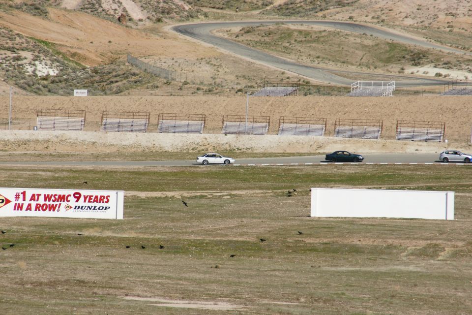 Race track with three cars driving, a sign that says "#1 AT WSMC 9 YEARS IN A ROW! DUNLOP", and a white barrier