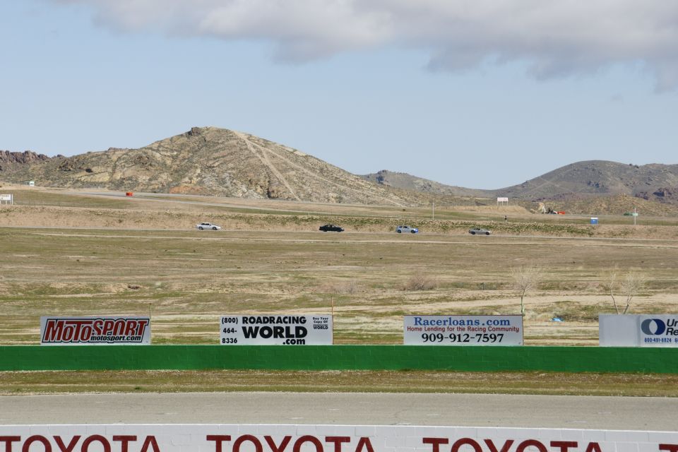 Several cars drive on a race track with dry grass and hills under a cloudy sky