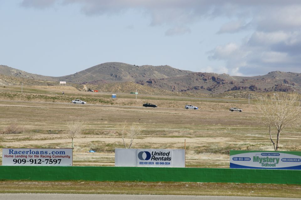 Several cars drive on a track with hills and signs in the background