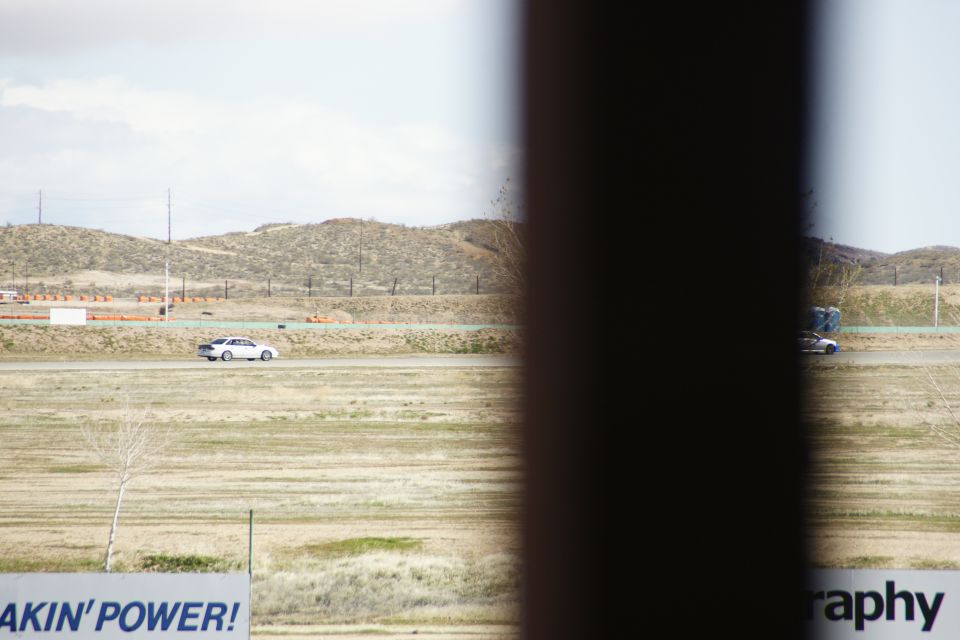 Two cars drive on a track with hills in the background. A dark pole is in the foreground