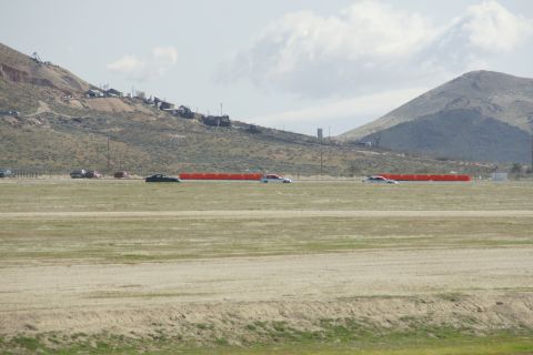 1995 Ford Taurus - Several vehicles are visible on a track, with orange barriers and a distant hillside settlement under a cloudy sky.
