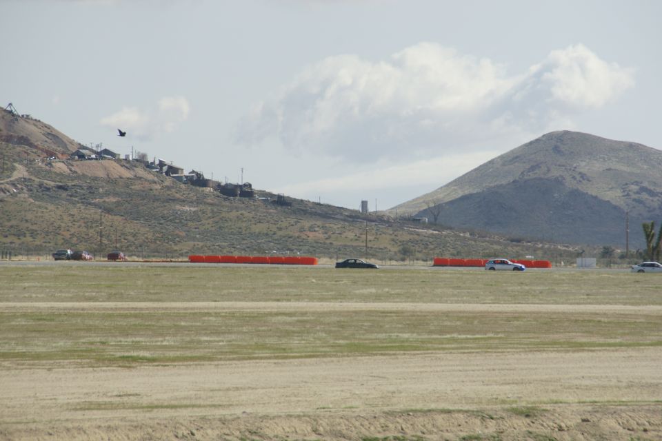 Cars driving on a track with orange barriers, hills, and buildings in background