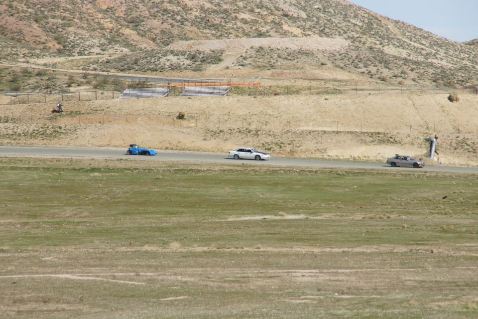 Three cars race on a track with dry, hilly terrain in the background