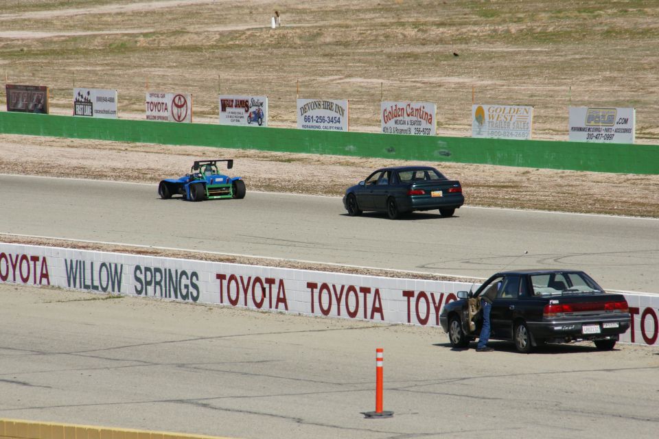 Three vehicles on a race track, including a dark sedan and a blue and green open-wheel car