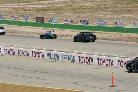 1995 Ford Taurus - A dark sedan drives on a race track alongside a blue open-wheel car, with 'Willow Springs' visible on the wall.