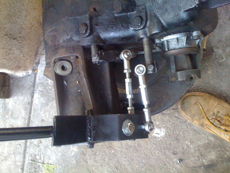 Transfer case linkage with adjustable rods and a black bracket. A tan boot is visible on the right