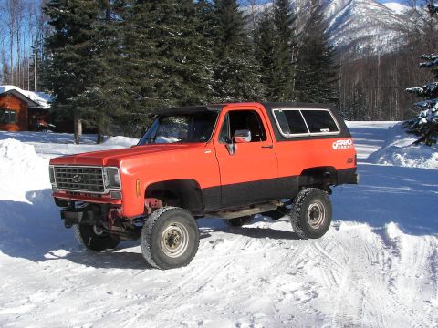 1990 Chevrolet Suburban - This lifted SUV, likely from the 1970s, is parked in a snowy landscape with trees and mountains in the background.