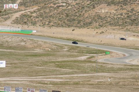 1995 Ford Taurus - Two dark sedans navigate a sweeping turn on a sun-drenched racetrack, with distant hills and advertising in the background.