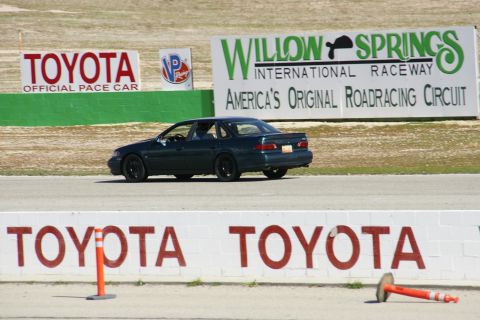 1995 Ford Taurus - A dark 1995 Ford Taurus sedan drives on a racetrack with Willow Springs International Raceway signage in the background.