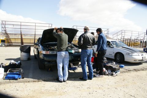 1995 Ford Taurus - People work on a dark green sedan with its hood open, while a white sedan with racing stripes sits nearby.