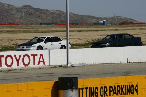 1995 Ford Taurus - Two sedans race on a track, one white with a black hood and the other dark green, with a white barrier and 'TOYOTA' logo visible.