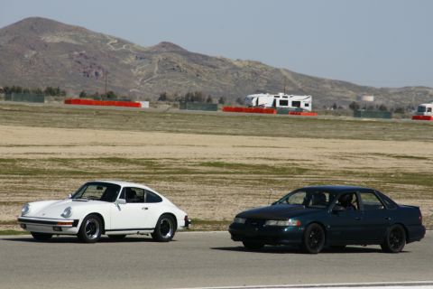 1995 Ford Taurus - A white Porsche 911 and a dark green sedan are on a racetrack, with mountains and RVs in the background.