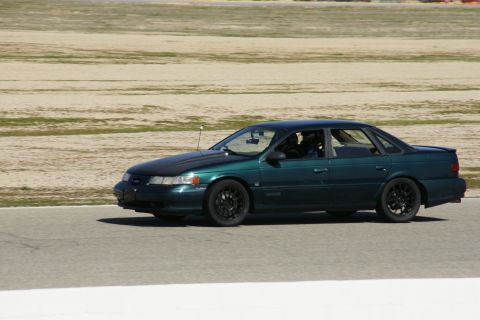 1995 Ford Taurus - A green sedan with a black hood and black wheels drives on a paved track with a dirt infield.