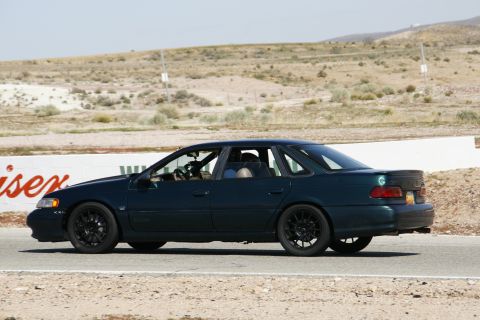 1995 Ford Taurus - A dark green sedan is captured in profile on a race track, its black rims contrasting with the body.