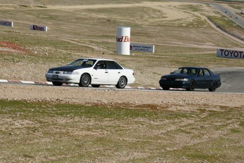 1995 Ford Taurus - Two sedans, one white and one dark, driving on a race track with tire barriers.