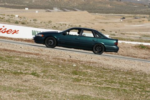 1995 Ford Taurus - A dark green sedan is captured in profile as it navigates a turn on a paved track with dry, grassy hills in the background.