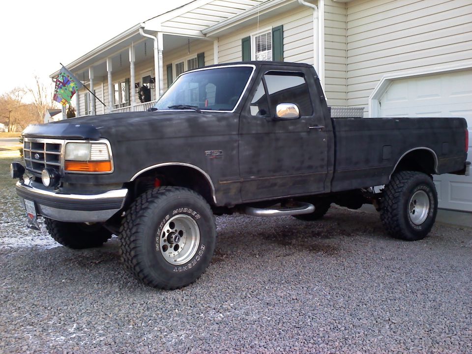 Lifted flat black Ford F-150 regular cab pickup with Maxxis Mudder tires parked on gravel by a white house